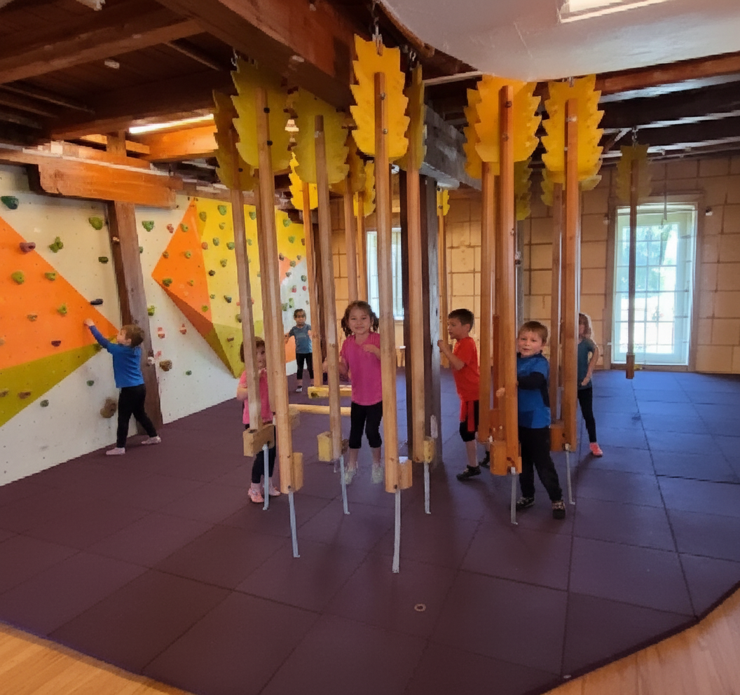 Children play in an indoor climbing room with a climbing wall and hanging elements.