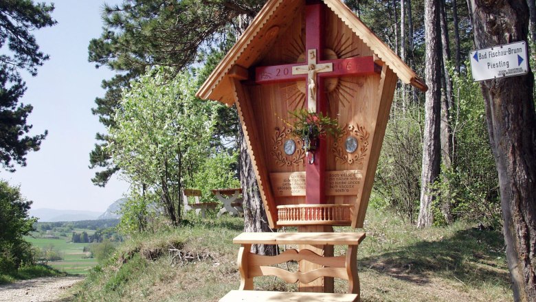 Wooden cross with red beam and carved roof in a forest landscape.