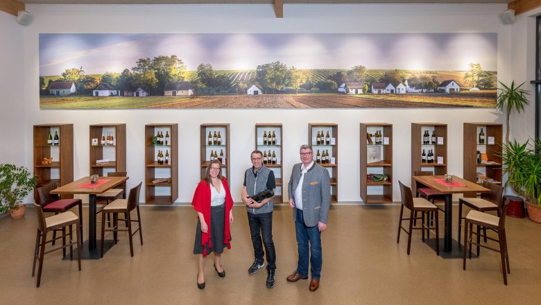 Three people are standing in a winery in front of shelves of wine bottles and a large mural of vineyards.