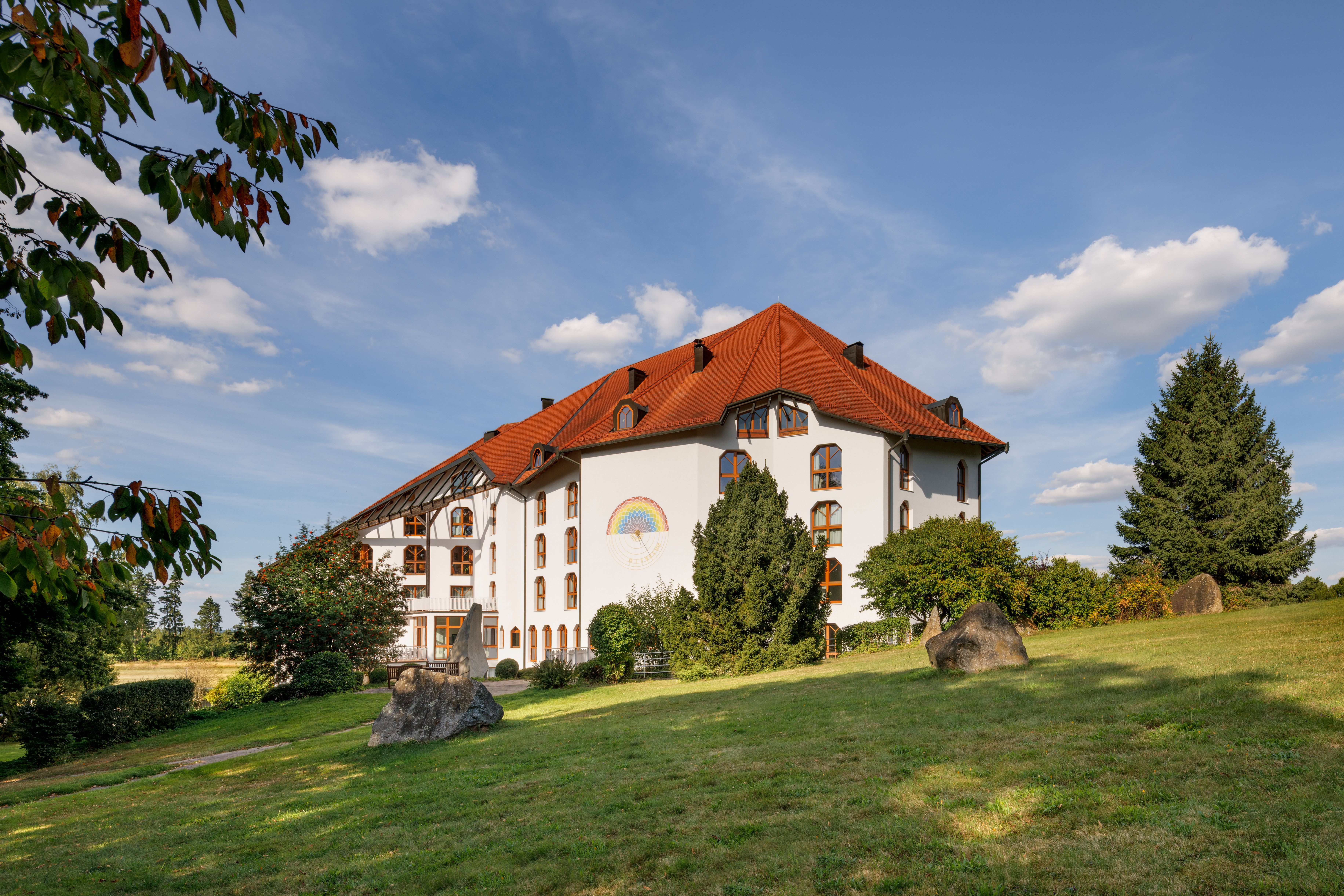 A large, white building with a red roof and a rainbow picture on the wall, surrounded by a green landscape and trees under a blue sky.
