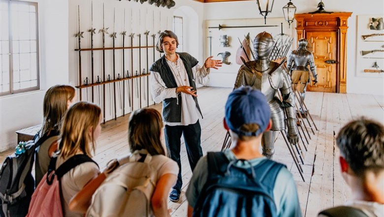 A guide explains a suit of armor to a group of visitors in a room full of medieval weapons at Rosenburg Castle.