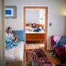 Interior view of a vacation apartment with two rooms. In the foreground a girl is sitting on a bed, in the background two people are reading on another bed.