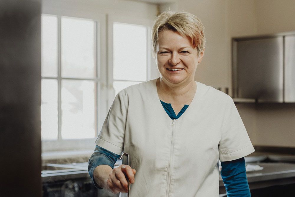 A smiling woman in a white chef's jacket stands in a kitchen.