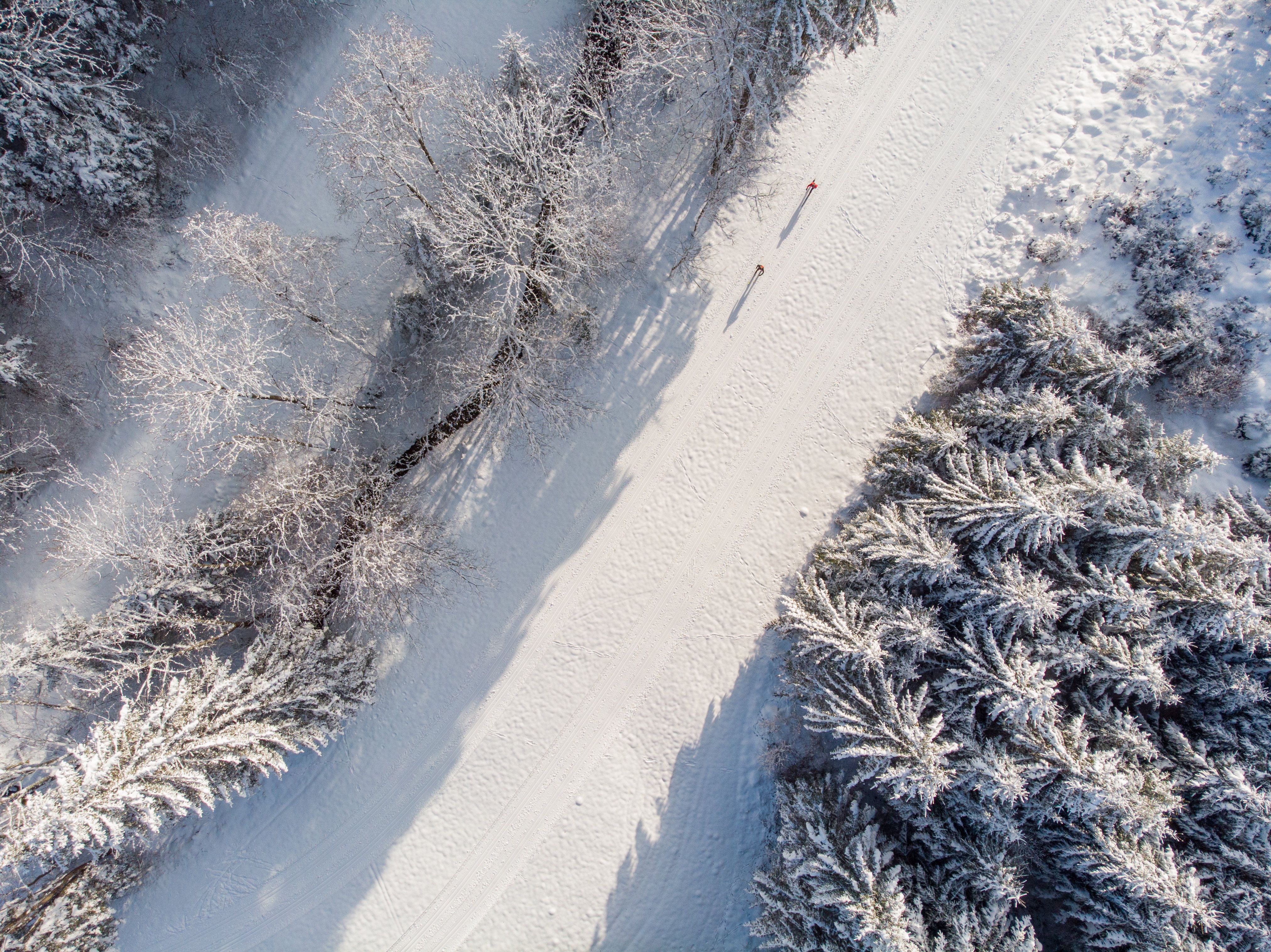 Aerial view of a snow-covered cross-country ski trail next to a stream with two people cross-country skiing, surrounded by snow-covered trees.