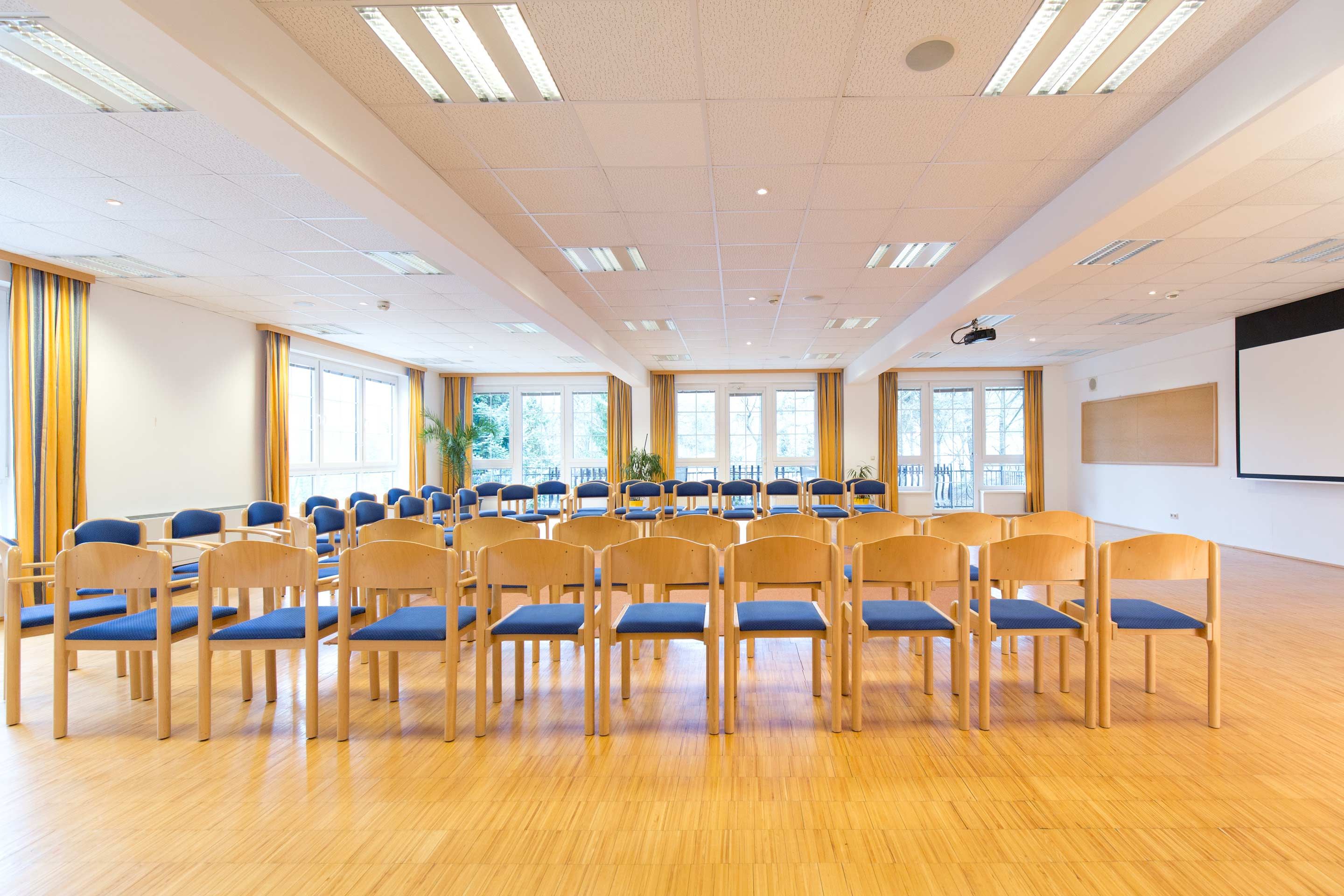 A bright seminar room with rows of chairs, large windows, and a projector mounted on the ceiling.