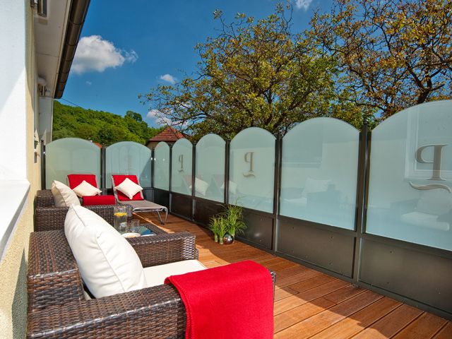 A terrace with rattan furniture, red blankets and cushions, surrounded by glass walls and trees in the background.
