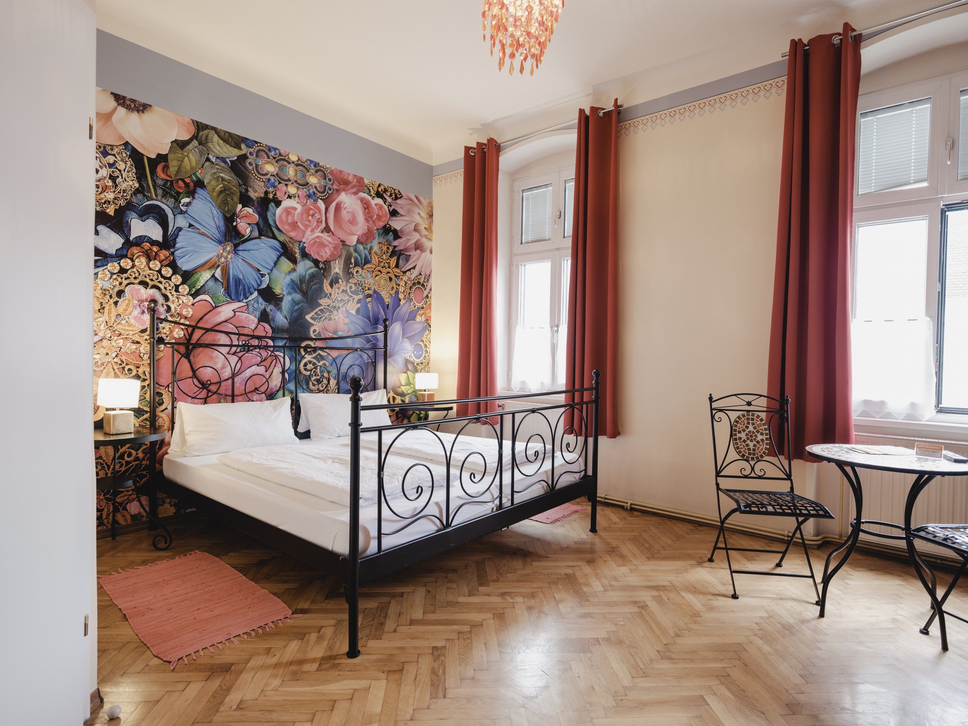 A stylishly furnished room with a wrought-iron bed, floral wallpaper and a small table with chairs in front of large windows.