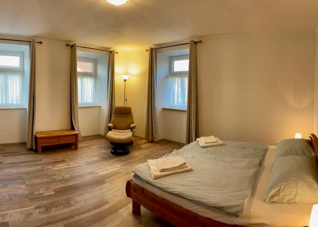 A simple bedroom with double bed, armchair, bedside table and three windows with curtains.