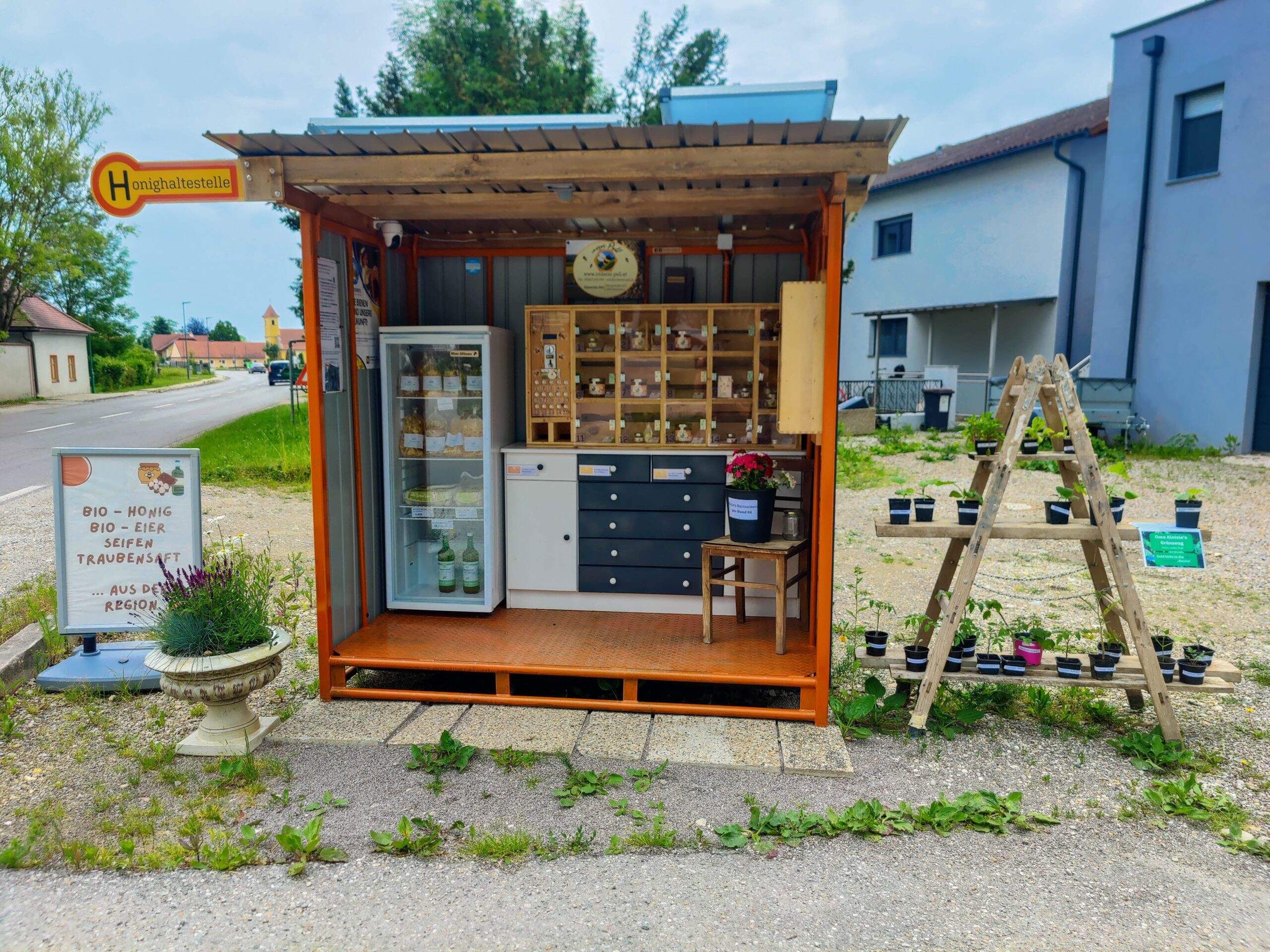 A small roadside stall selling organic products such as honey, eggs and soaps. Next to it are plants on a wooden frame.