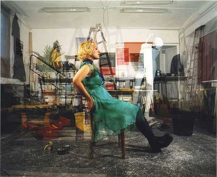 A woman in a green dress sits on a chair in a crowded room.