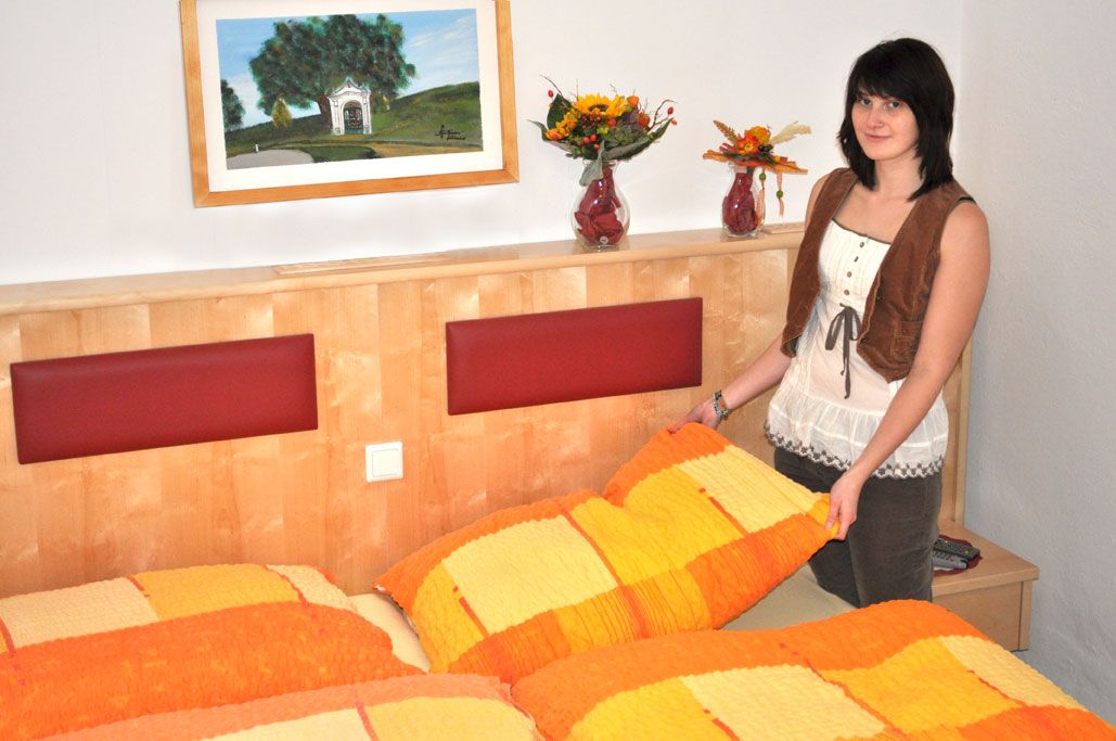 A woman stands in a bedroom with orange and yellow bed linen and vases of flowers on the headboard.