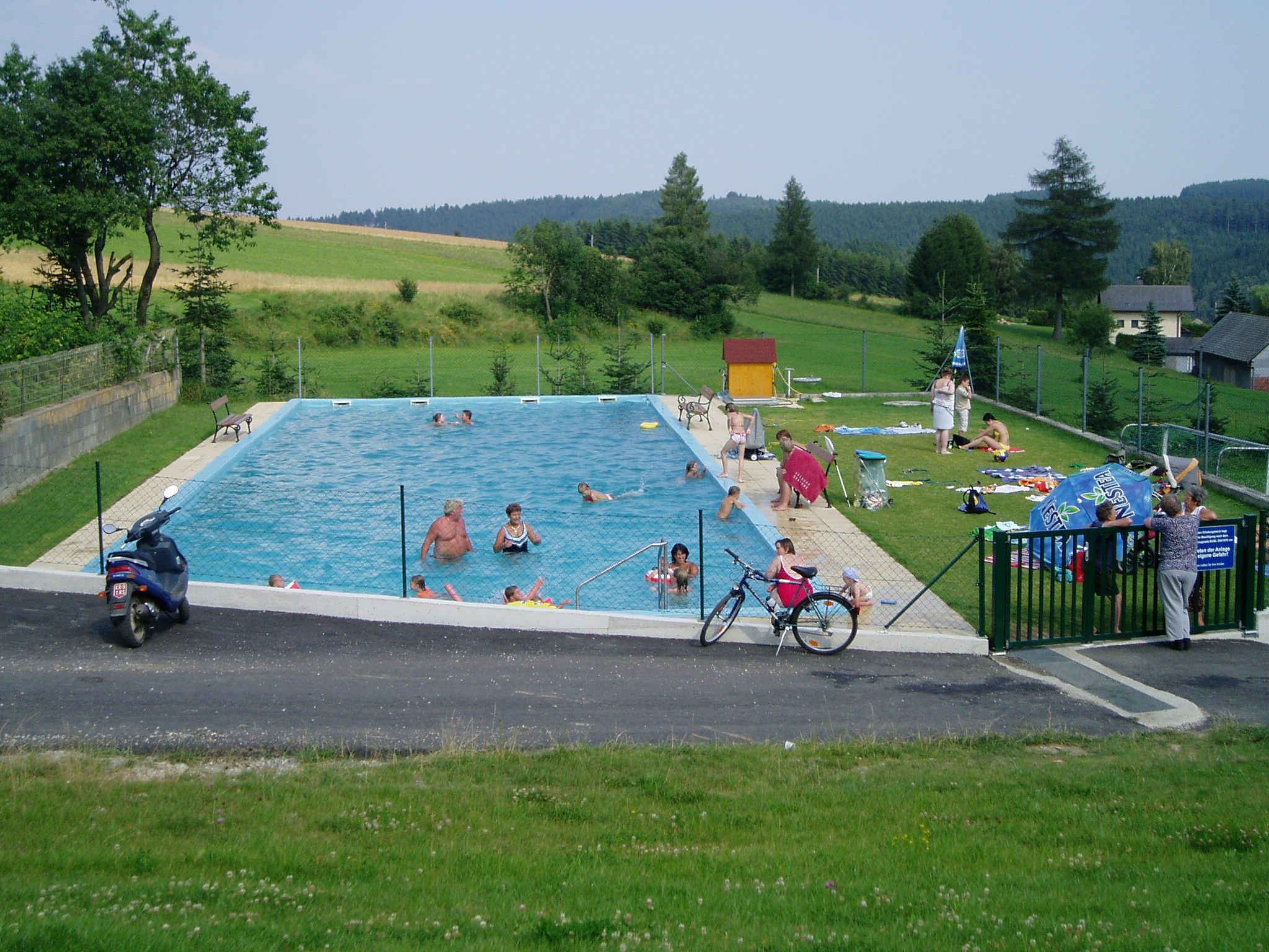An outdoor pool with several people, surrounded by green landscape and trees.