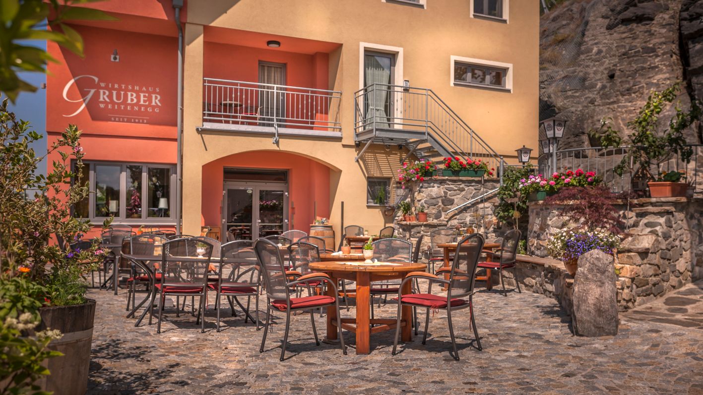Outdoor area of a restaurant with tables and chairs on a paved courtyard, surrounded by flowers and plants.