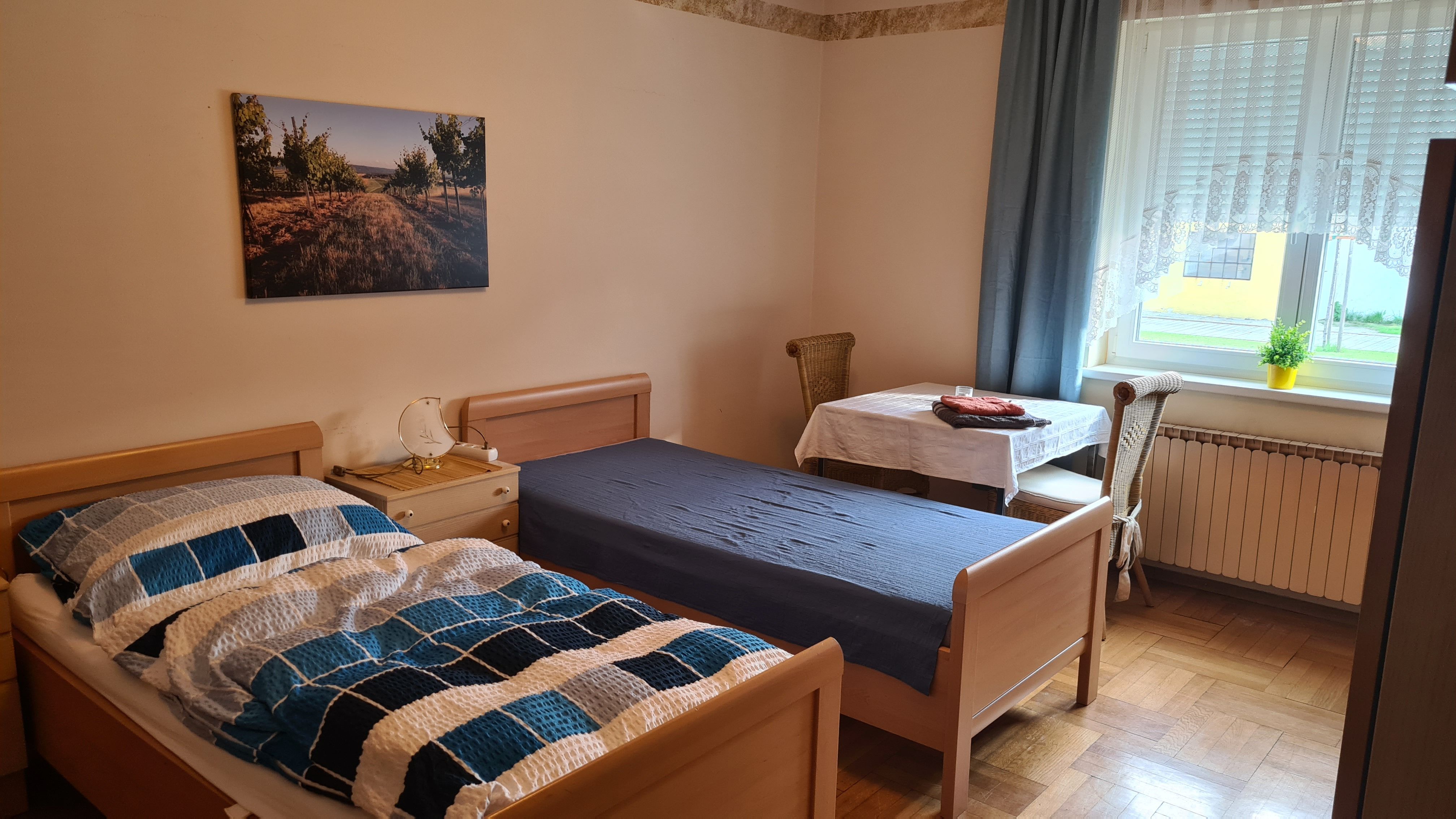 A simply furnished bedroom with two single beds, a table and chair and a window with curtains.