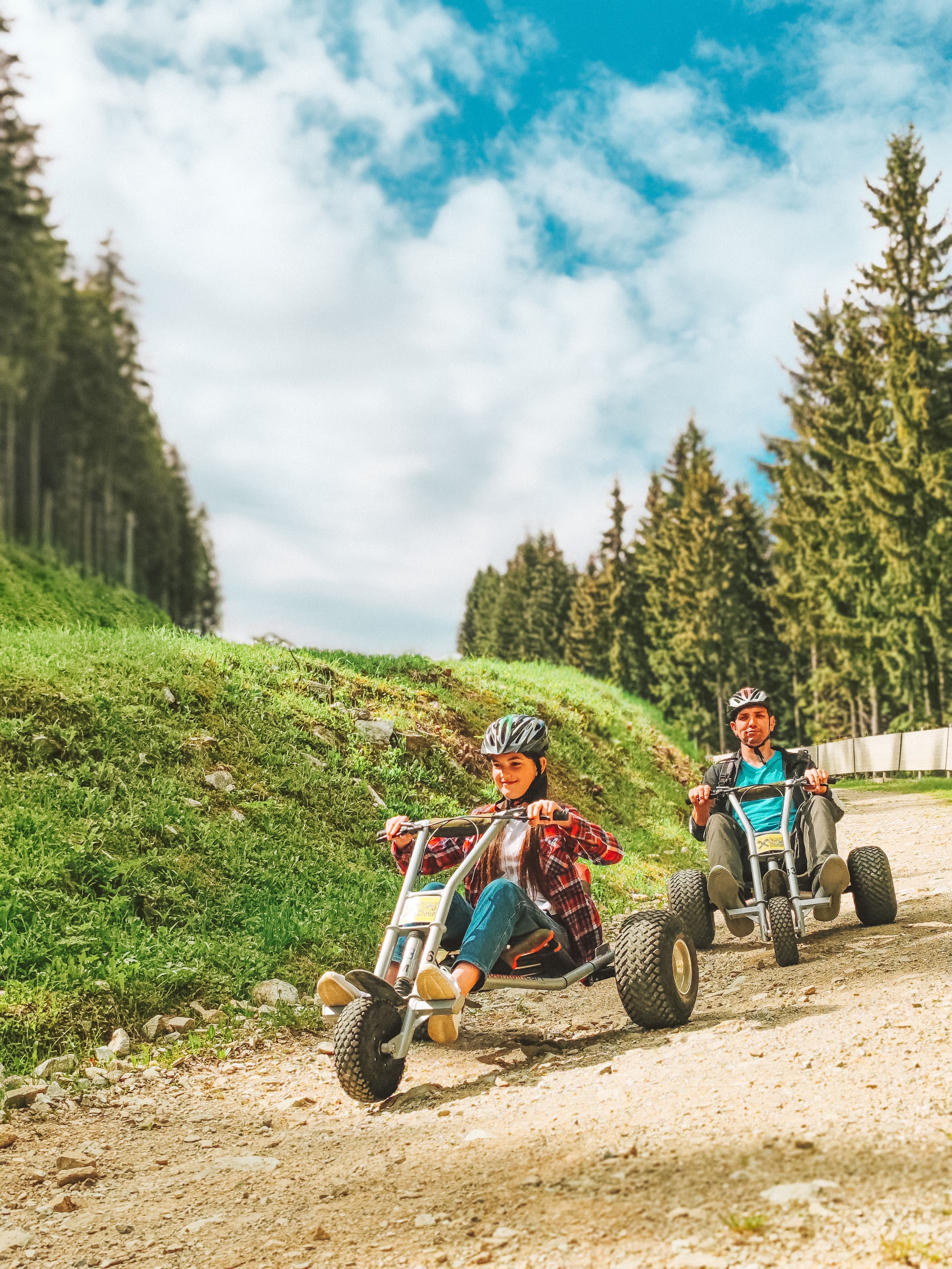 Two people ride mountain carts on a gravel road, surrounded by trees and blue sky.