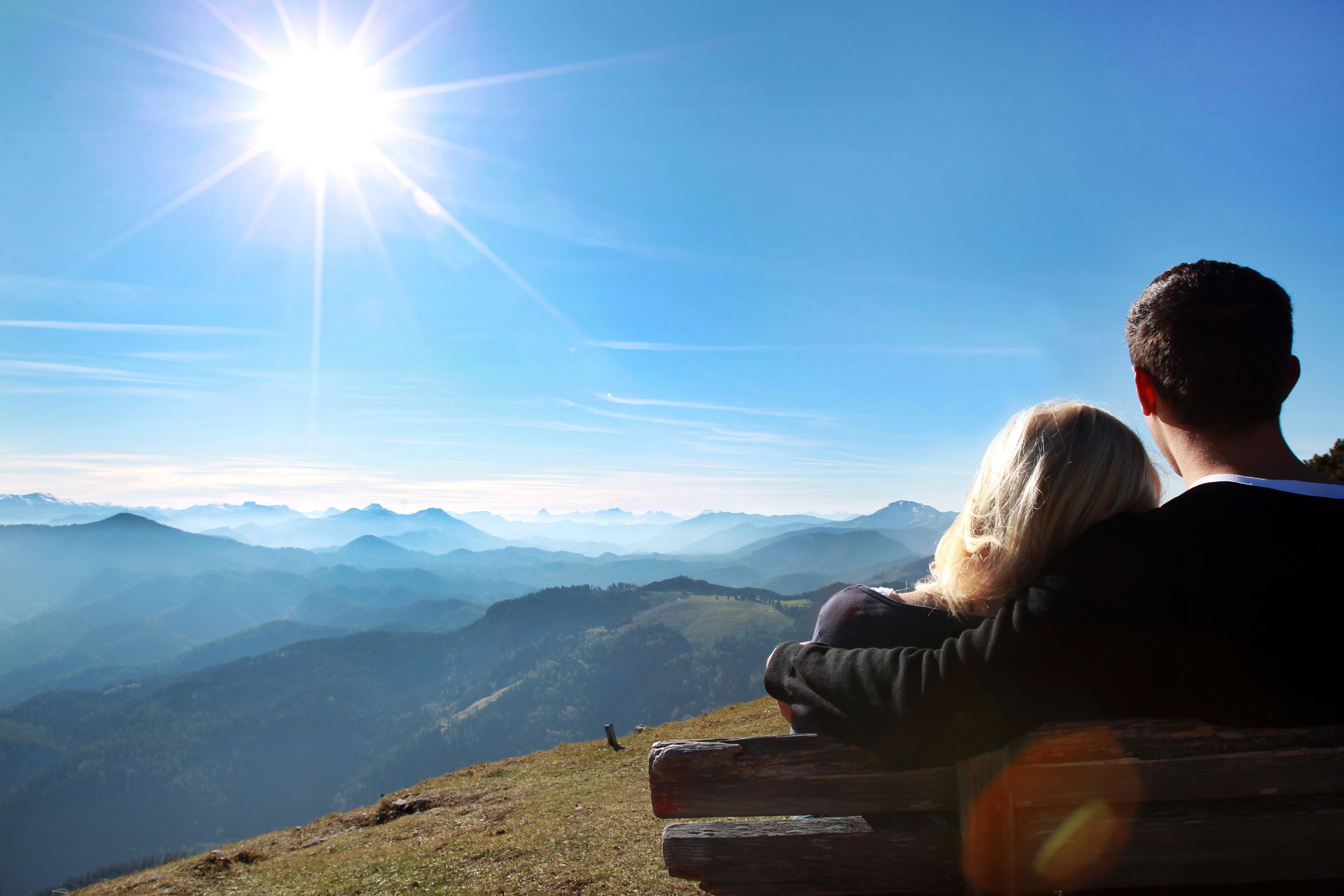 A couple sits on a bench and looks out over a mountain landscape under a bright blue sky and sun.