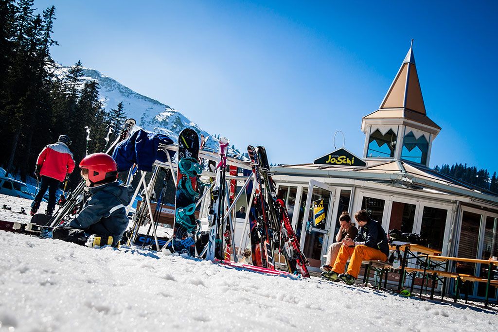 Skiers in front of the JoSchi bar with skis and snowboards in the snow.
