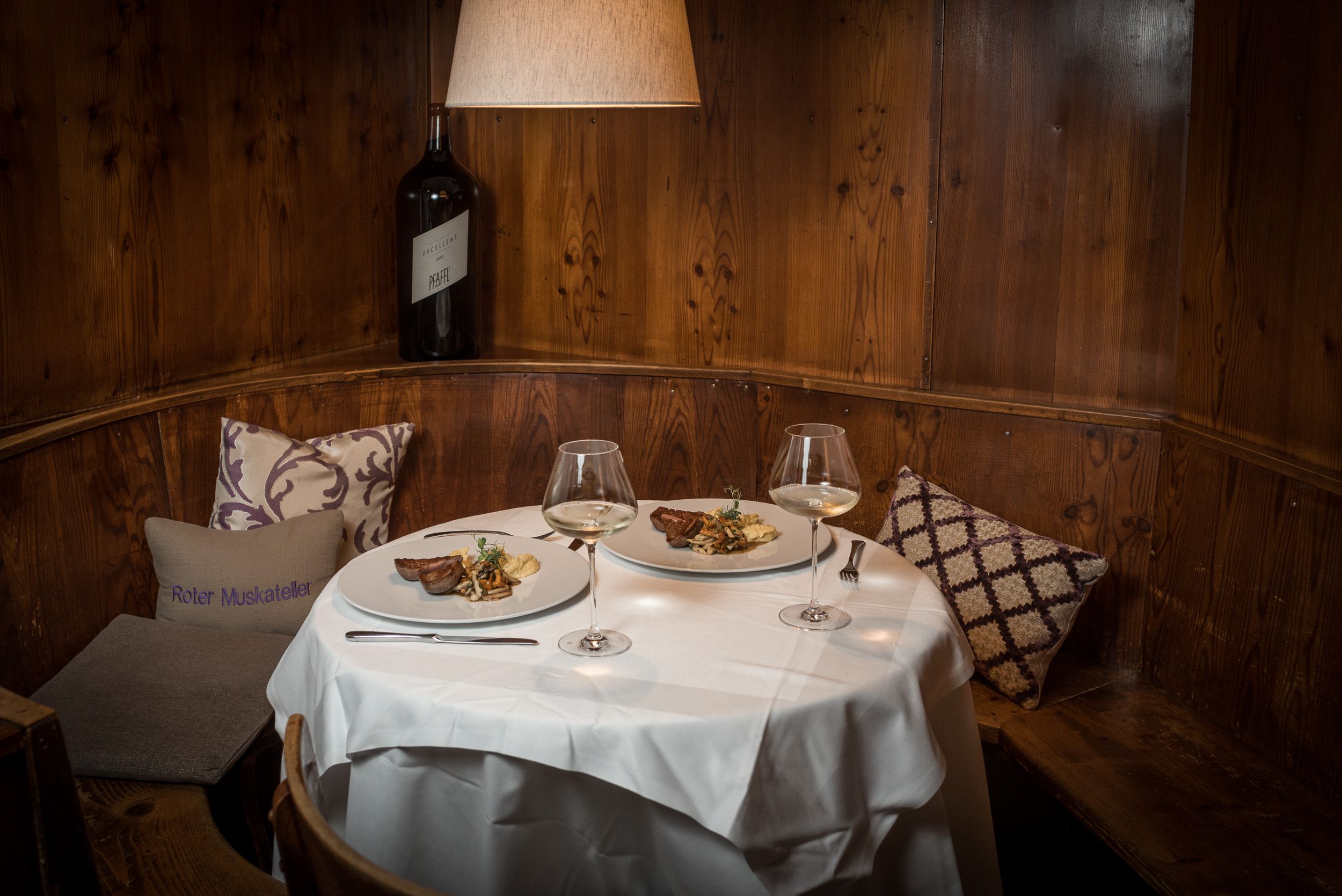 Cozy dining room with set table, two plates with food and wine glasses, wooden wall and cushions.