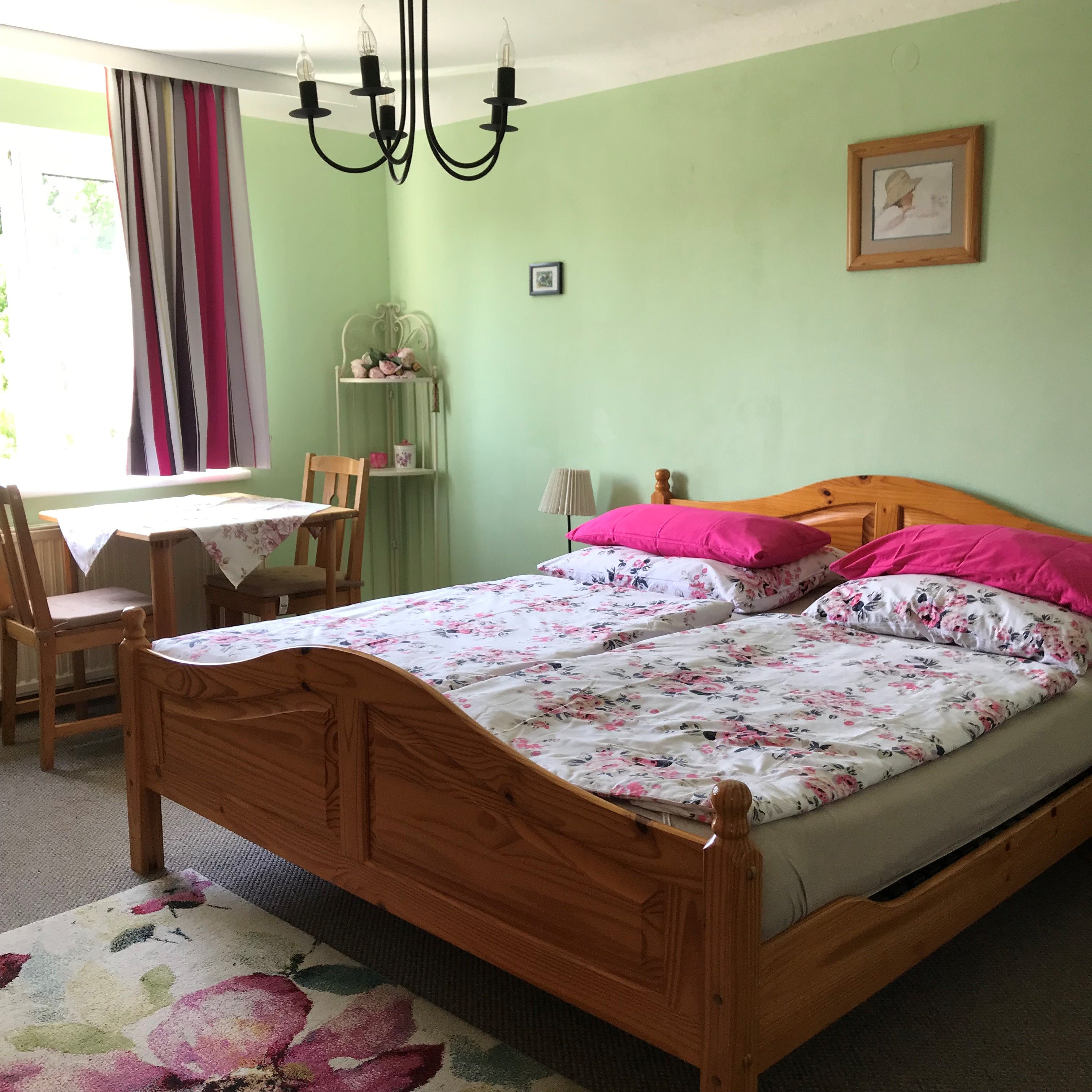 Bedroom with double bed, green walls, floral bedding, table with chairs, curtains and chandelier.