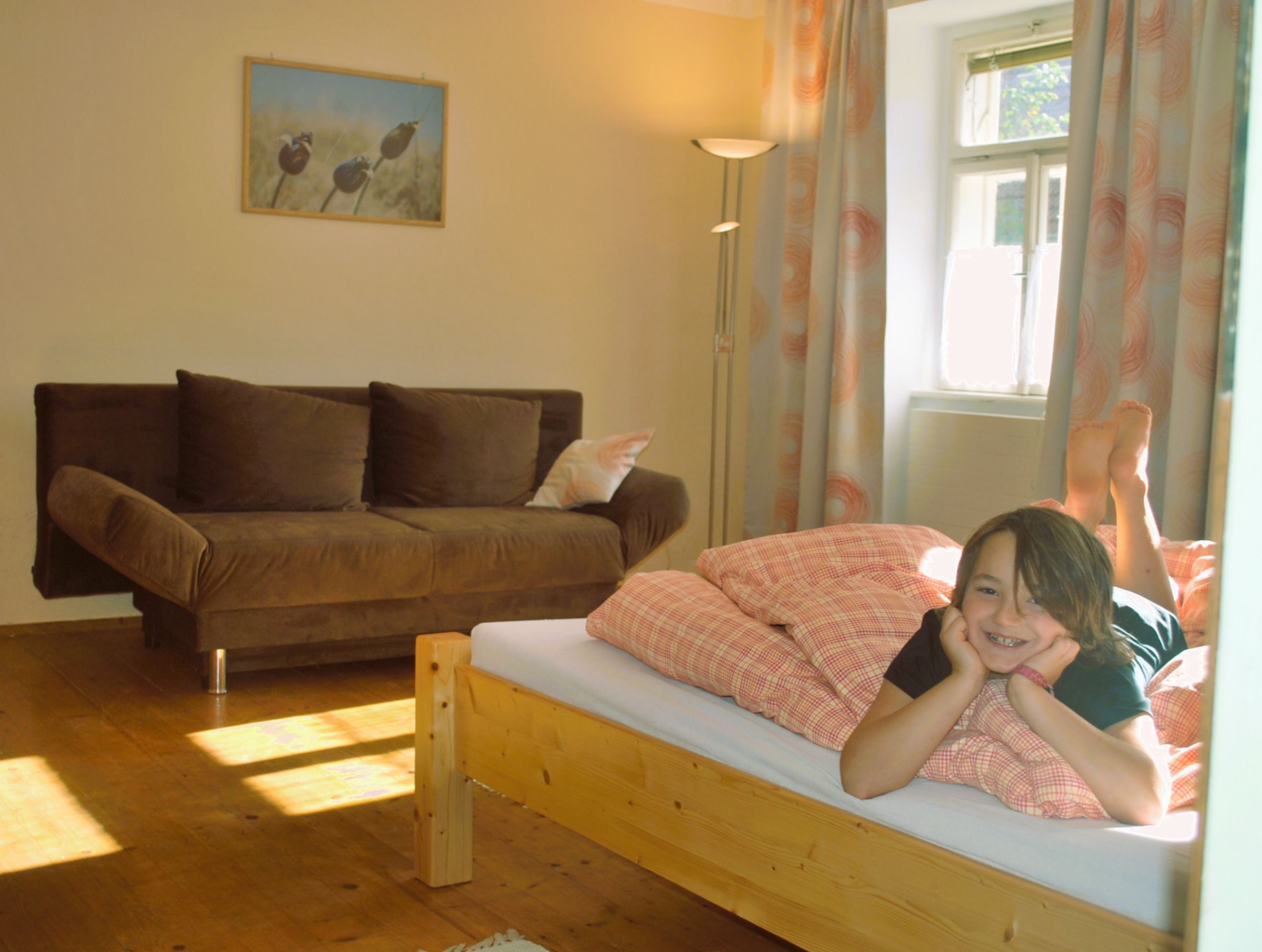 A child lies smiling on a bed in a cozy room with a sofa and a picture on the wall.