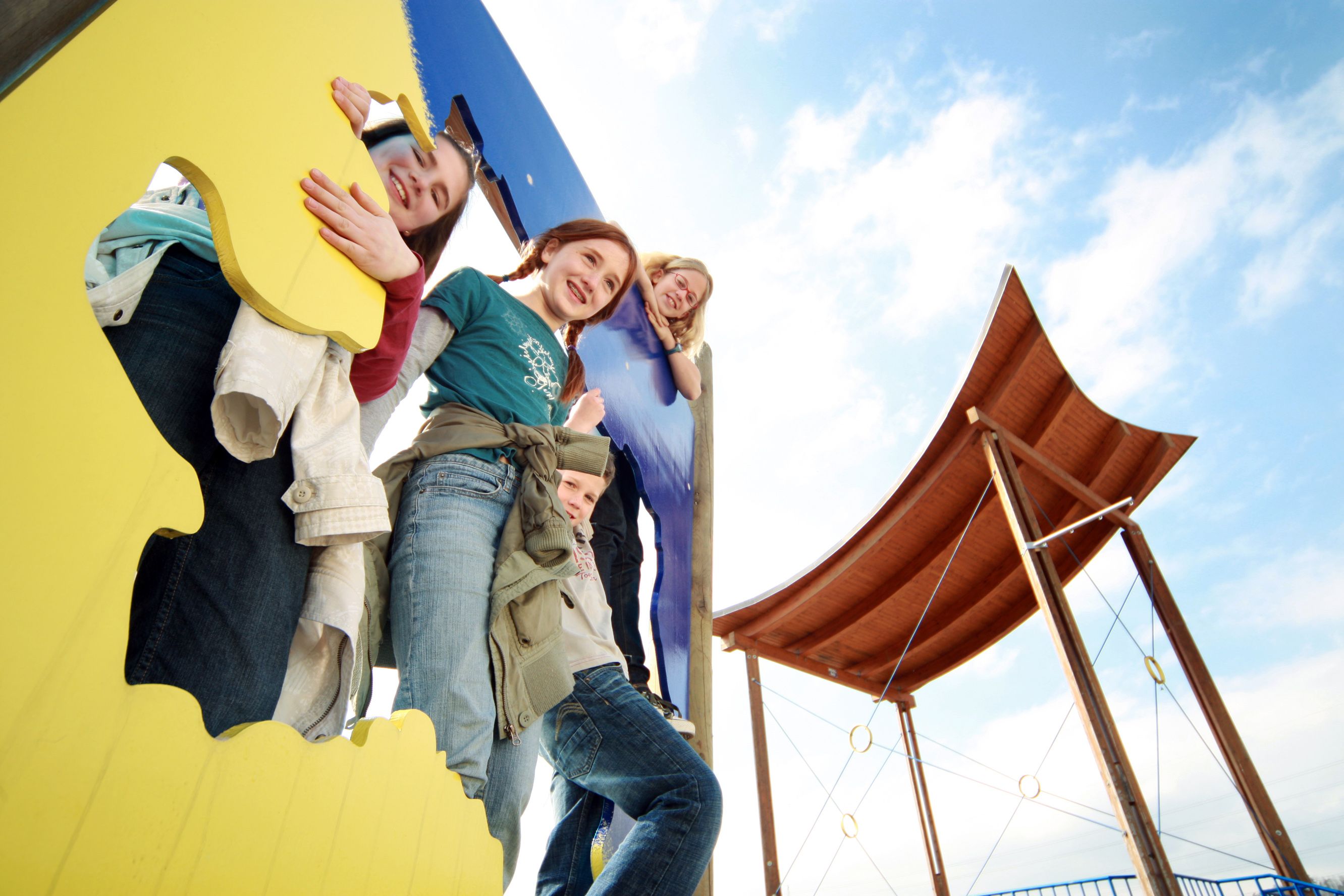 Children play on a colorful climbing frame with a wooden roof in the background.