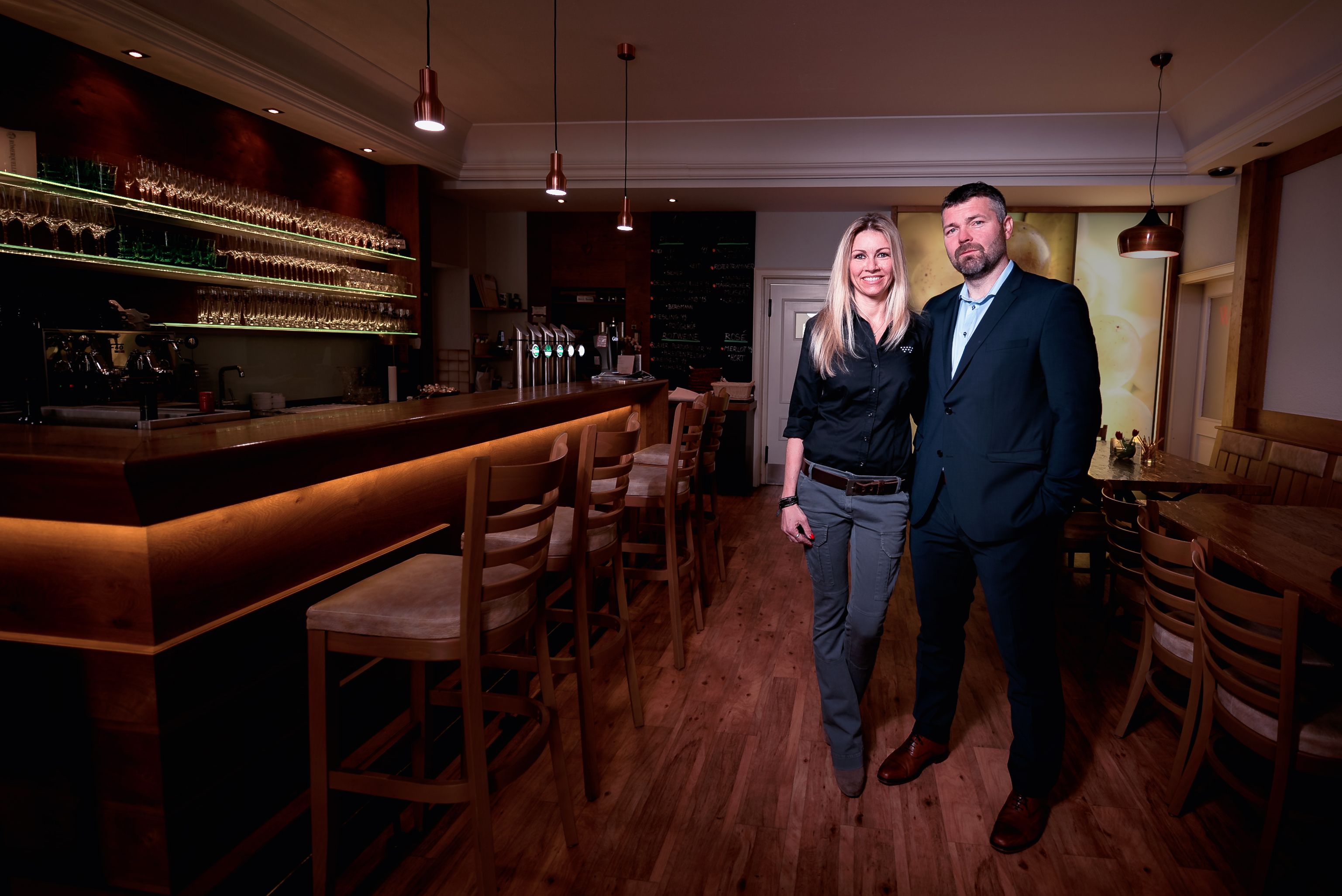 Two people stand in a stylish bar with a wooden counter and glass shelves.
