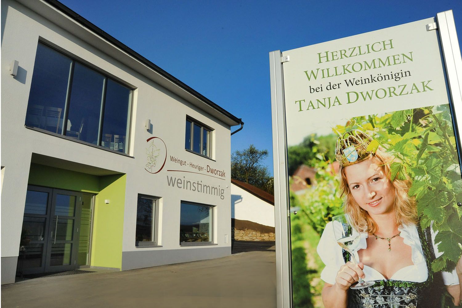 A modern building with a sign showing a woman in traditional costume with a wine glass. Text: Welcome to the wine queen Tanja Dworzak.