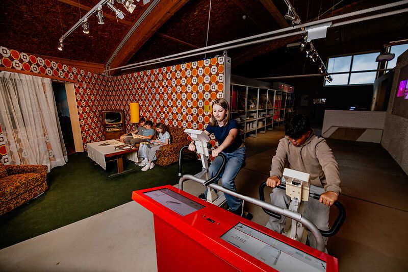 Young people on bicycle ergometers in a room with retro living room decor.