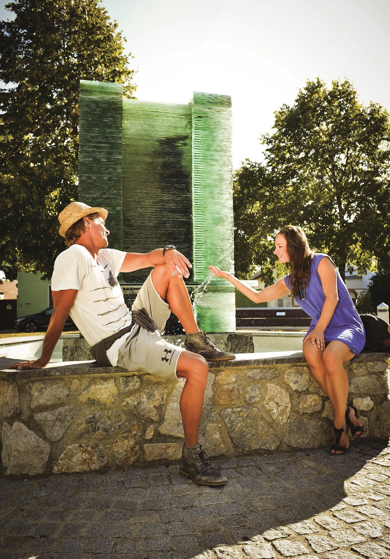 A couple is sitting on the wall in front of a fountain. The woman is playing with the water. 