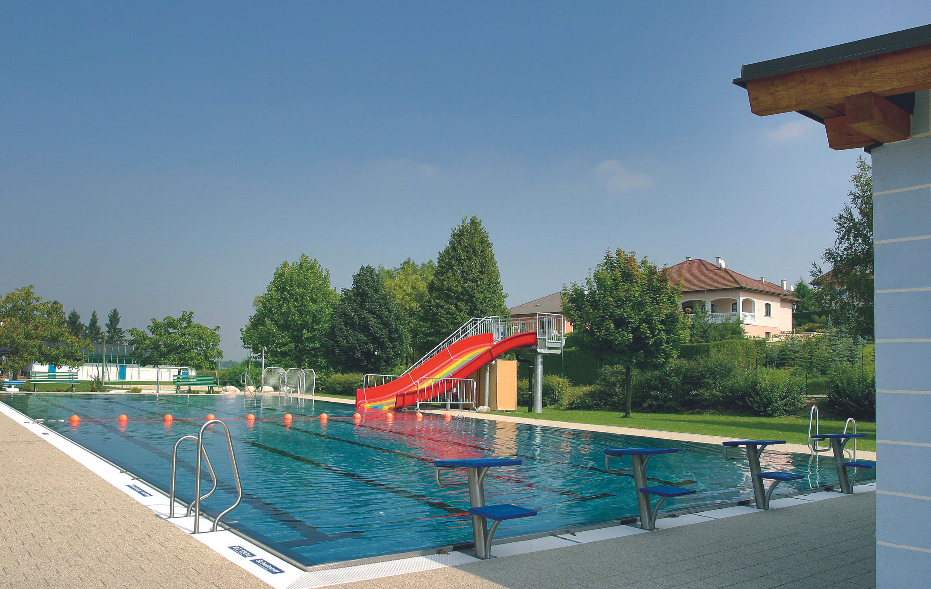 An outdoor pool with a swimming pool, a red slide and starting blocks. Trees and a house can be seen in the background.