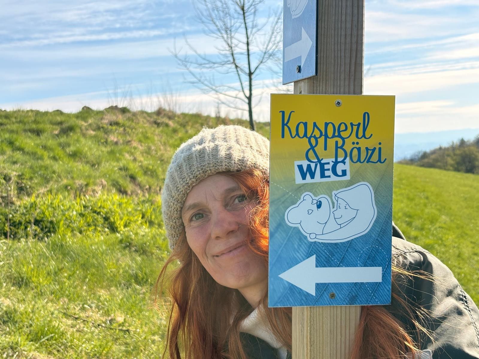 A woman wearing a cap is standing next to a sign that reads "Kasperl & Bäzi Trail" in a green landscape.