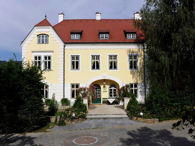 Yellow building with a red roof and green door, surrounded by trees and plants.