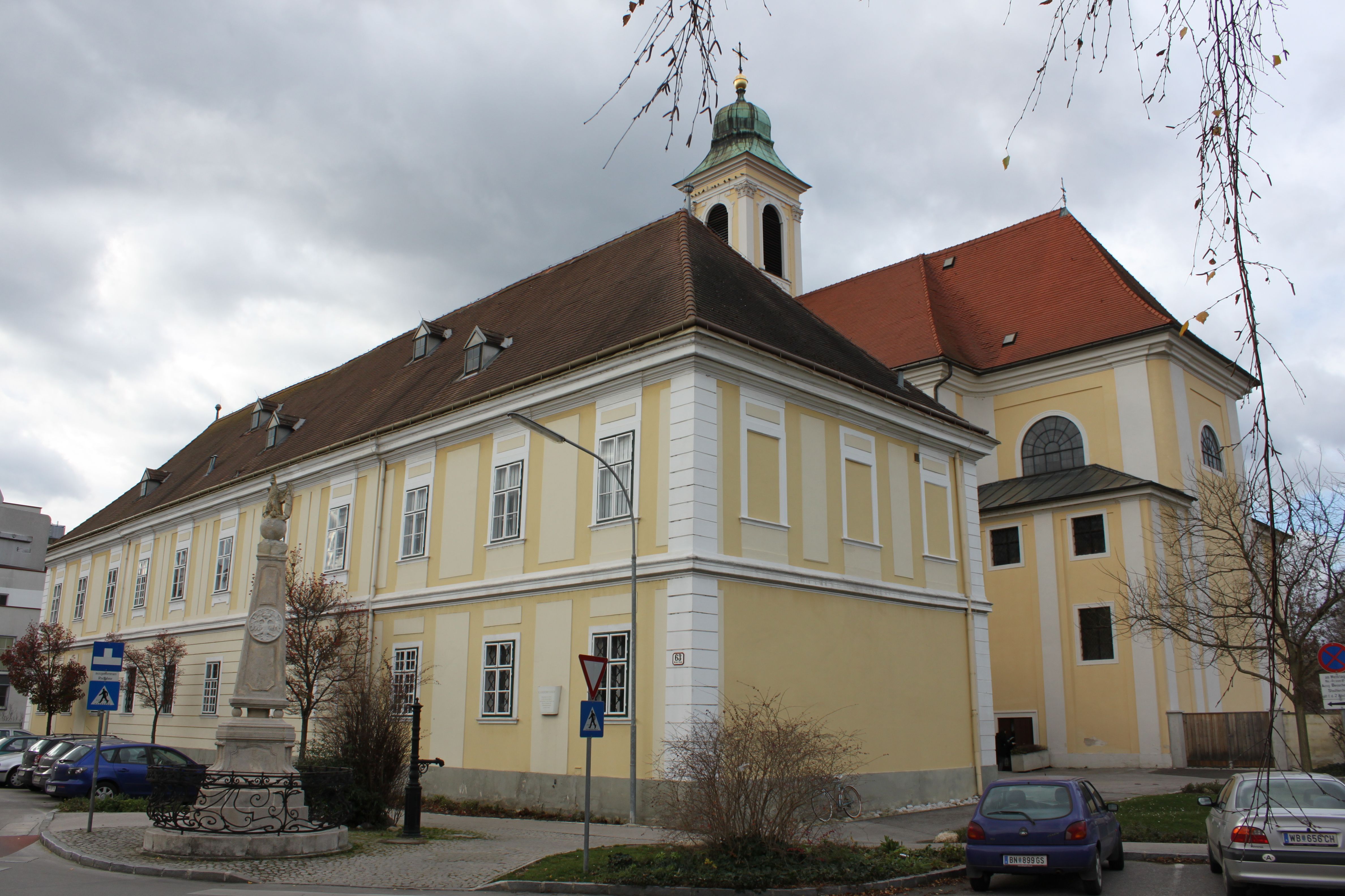 Historic building with a yellow façade and red roof, next to a statue and parked cars.