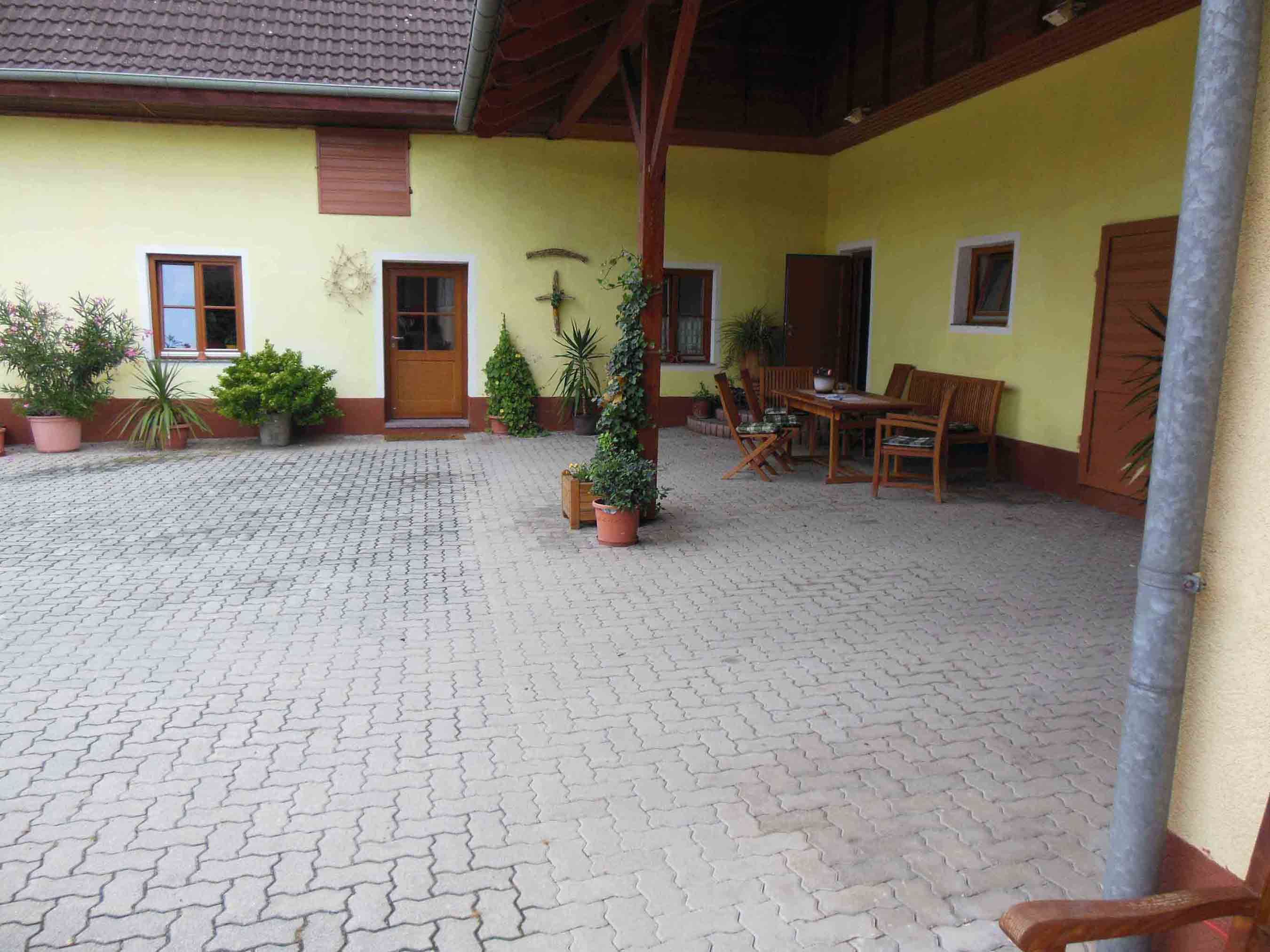 Inner courtyard with yellow façade, wooden table and chairs, surrounded by plants in pots.