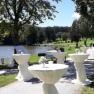 Bar tables with white cloths on the shore of a lake, surrounded by trees.