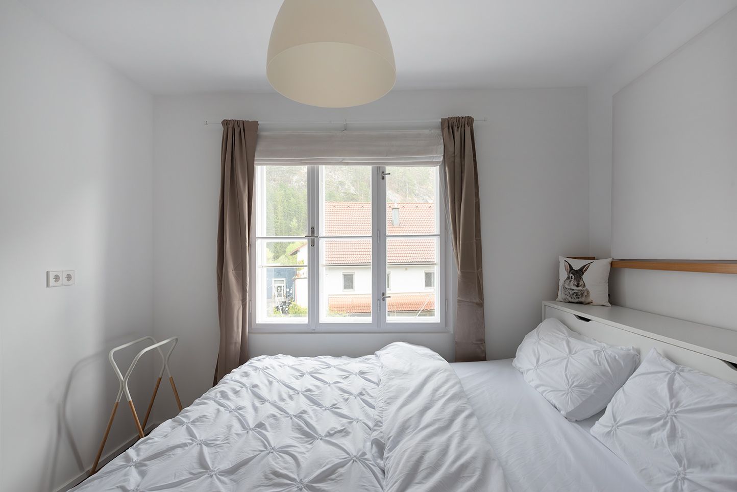 Bright bedroom with white bed and window overlooking a rooftop.