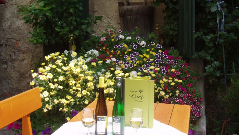 An outdoor table with bottles of wine, glasses and a menu, surrounded by colorful flowers.