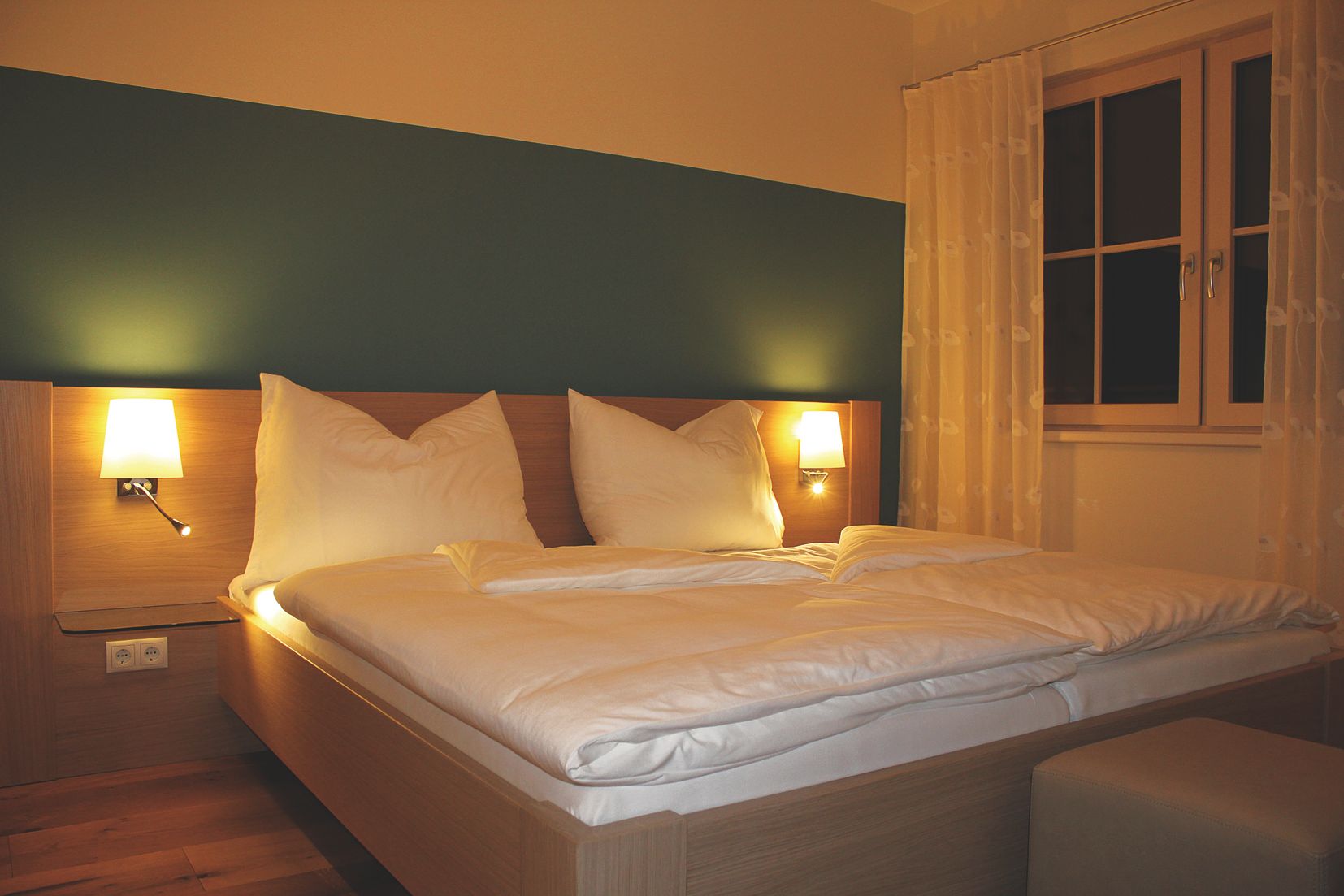 A cozy guest room with a double bed, two bedside lamps and a window with curtains.