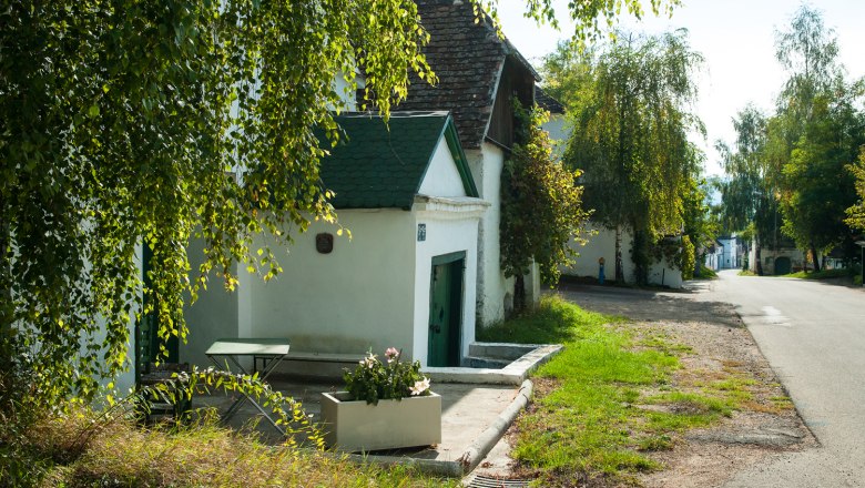 A quiet wine cellar lane with white buildings and green doors, surrounded by trees and plants.