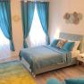 A cozy bedroom with blue curtains, a double bed and decorative cushions. Two bedside lamps illuminate the room.
