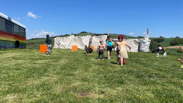 Children play on a meadow with archery targets and a colorful bus in the background.