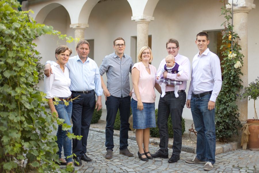 A family stands outside in front of a building with arches, surrounded by plants.