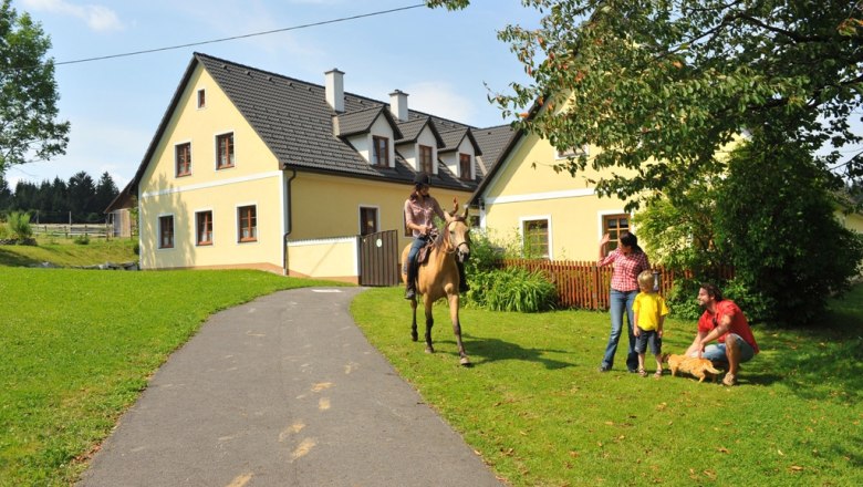 Hacklhof, © Urlaub am Bauernhof A rider on a horse approaches a family in front of a yellow house on a farm.