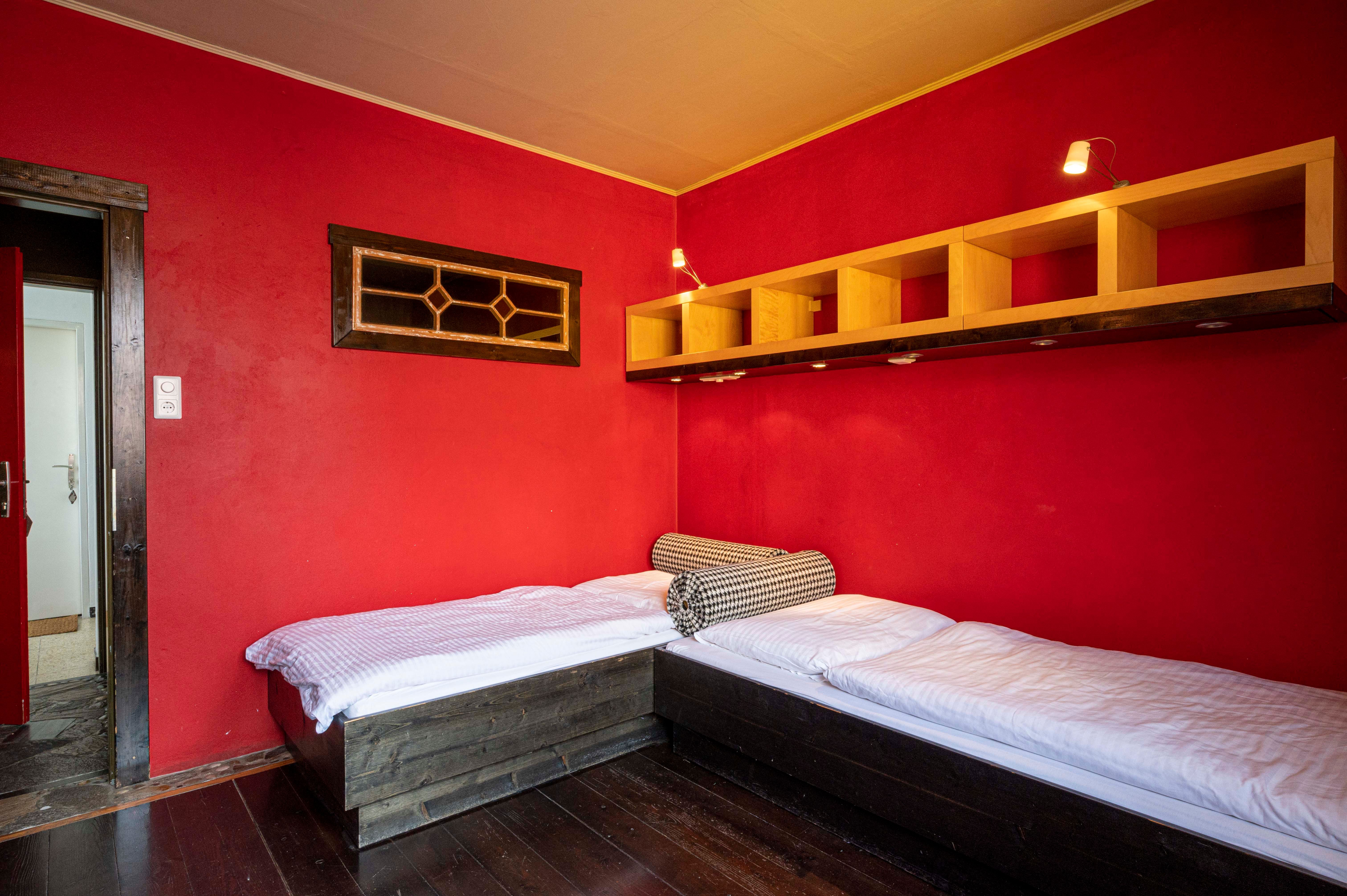 A room with red walls, two single beds and a shelf on the wall.