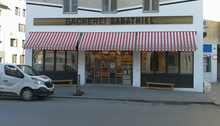 Exterior view of the Sabathiel bakery with red and white striped awning and delivery van in front.