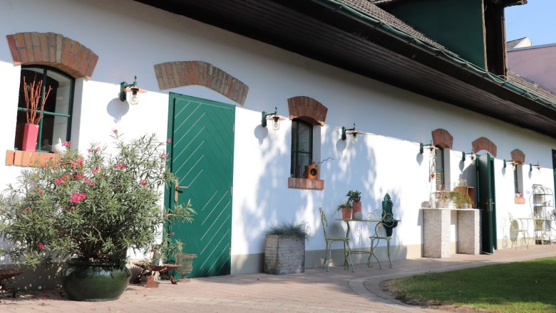 A white building with green doors and windows, surrounded by plants and garden furniture.