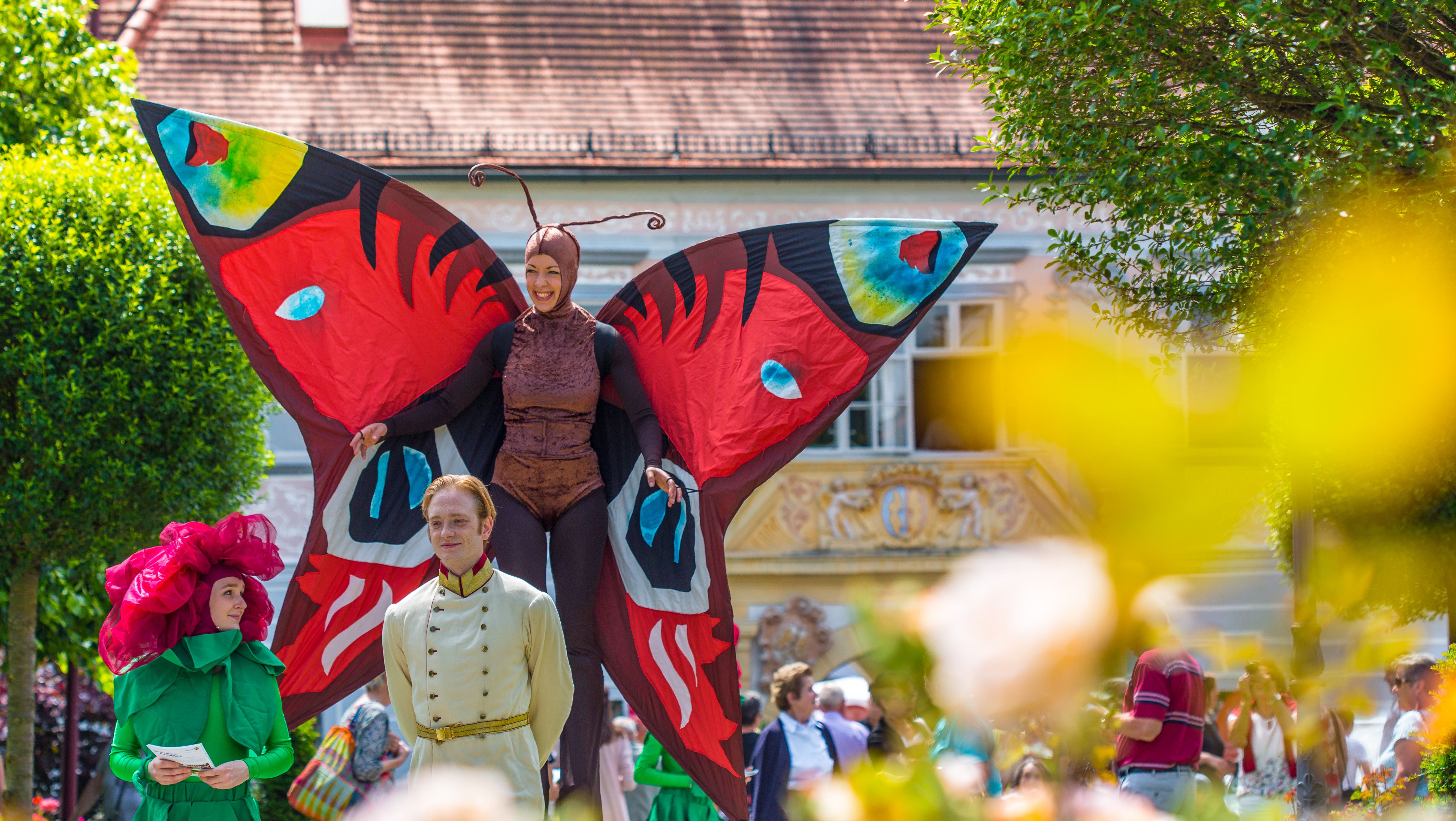 People dressed up in colorful costumes at the Rose Festival, one woman is dressed on stilts as a large colorful butterfly