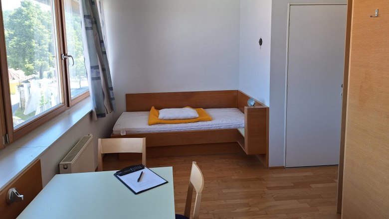 A simply furnished single room with a bed, table and chair, light wooden furniture and a large window with a view outside.