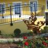 A yellow building with a balcony and a garden with red roses and a metal crown as decoration.