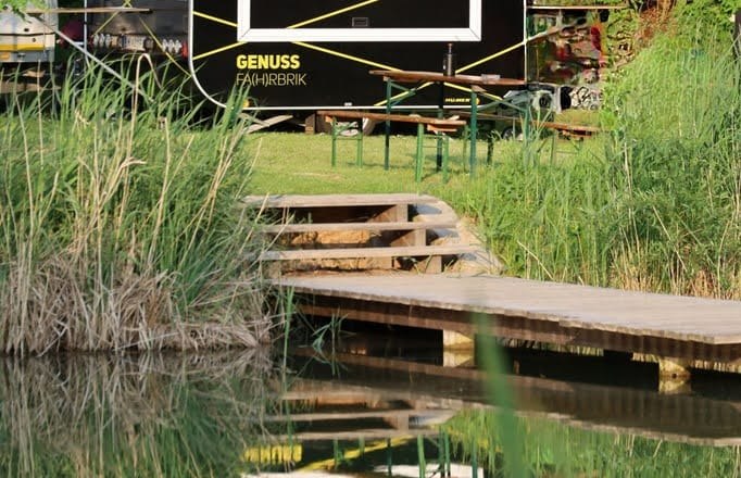 A black food truck with the inscription 'ZWOA Weinviertla' stands in a green setting, reflected in the water.